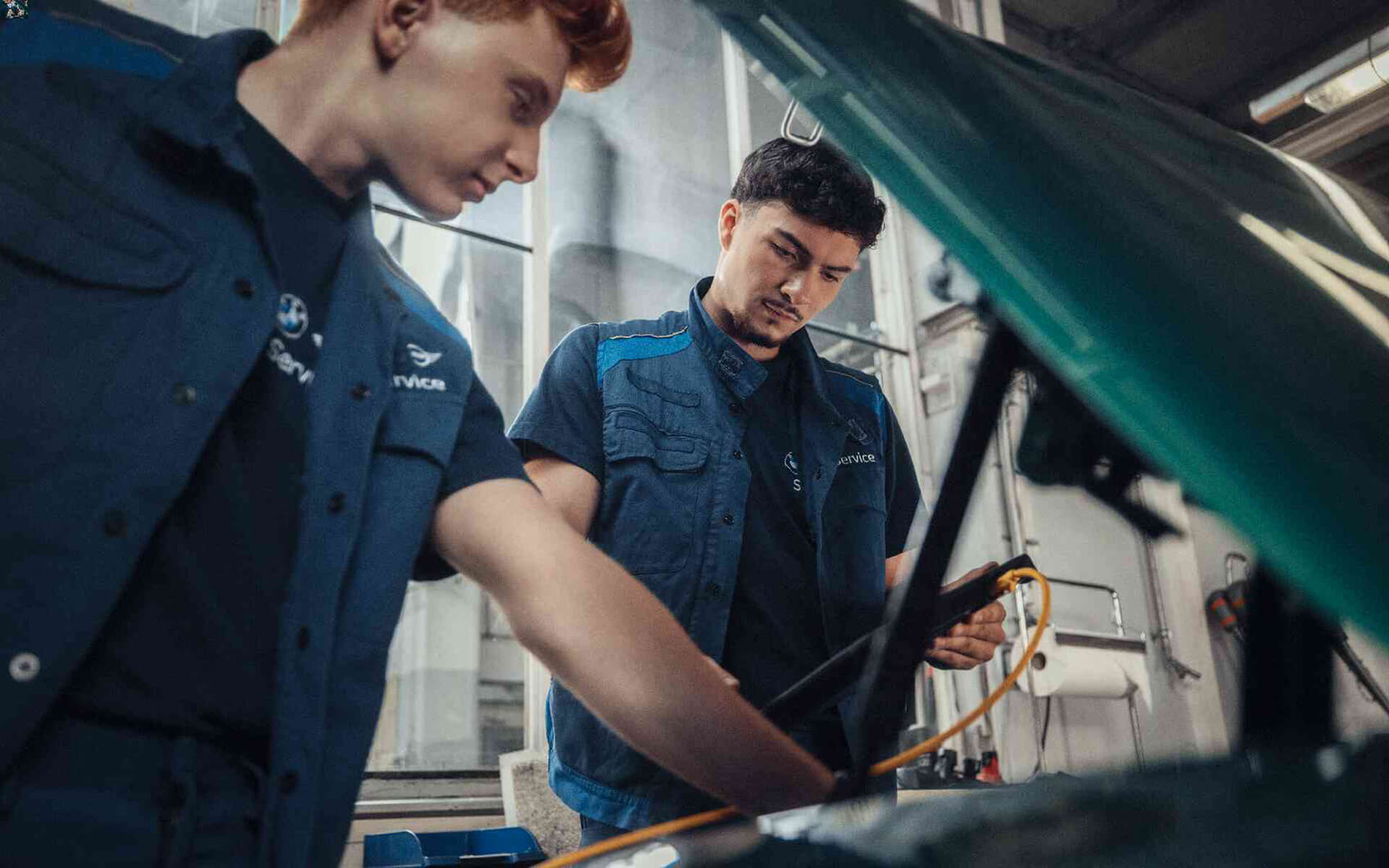 Zwei Kfz-Mechatroniker arbeiten gemeinsam am Motorraum eines Fahrzeugs.