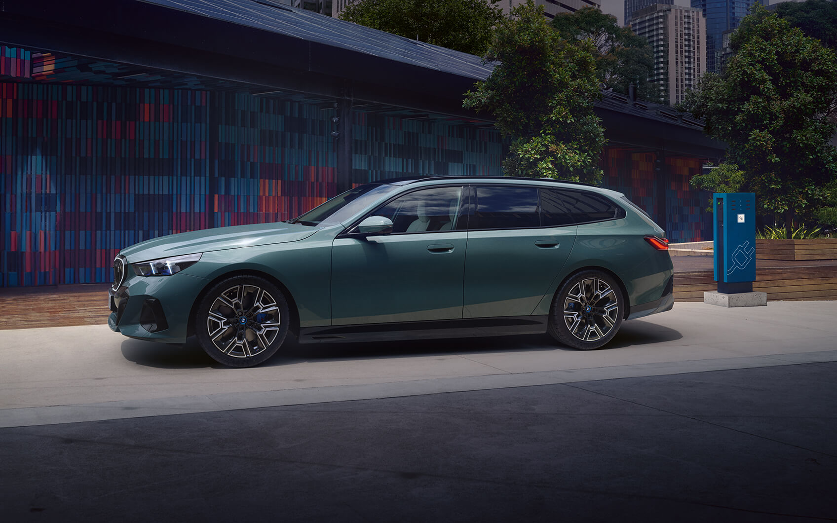 Grüner BMW i5 Touring an einer Ladestation in urbaner Umgebung.