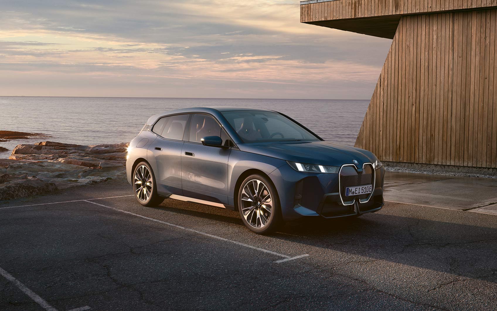BMW iX in der Farbe Blau rückwärts auf einem Parkplatz geparkt mit Meer im Hintergrund, Fokus auf die Beifahrerseite des Fahrzeugs