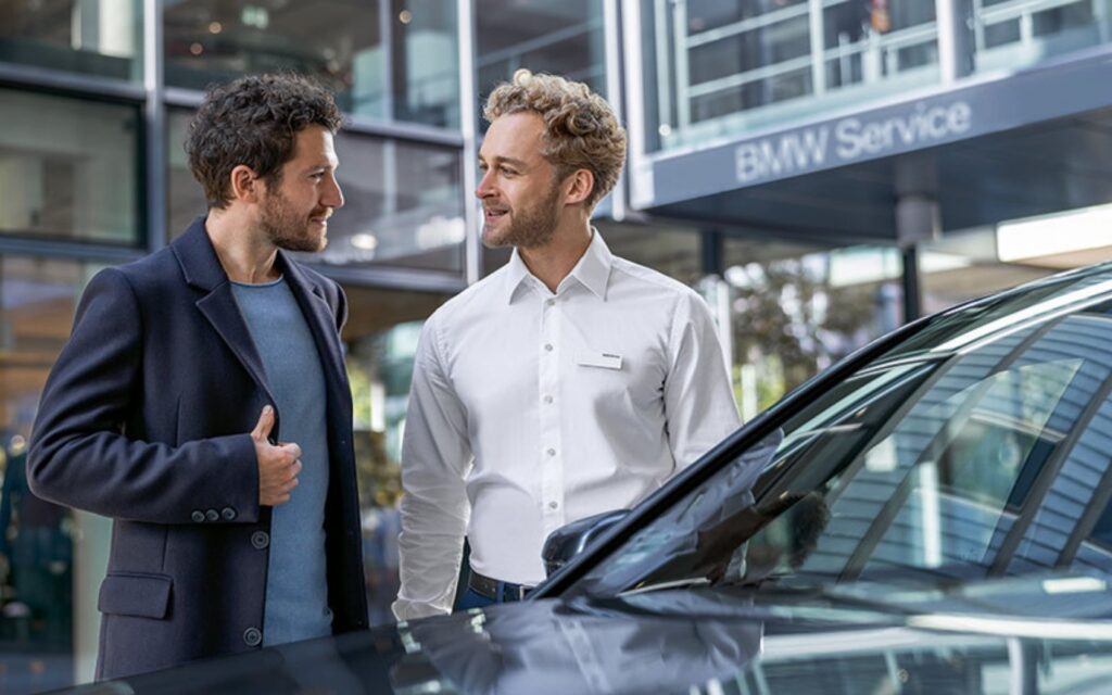 BMW Serviceberater im Gespräch mit einem Kunden neben einem Fahrzeug im Autohaus.