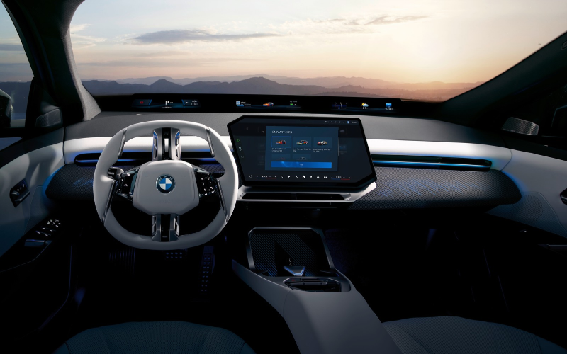 Blick ins Cockpit eines BMW mit Lenkrad, breitem Display und zentralem Touchscreen; durch die Windschutzscheibe ist eine Berglandschaft bei Sonnenuntergang zu sehen.